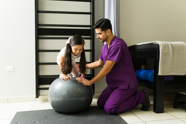 Profesionální fyzioterapeut pomáhá dětskému pacientovi vylézt na fitness ples a začít s fyzioterapeutickými cvičeními na klinice - Fotografie, Obrázek