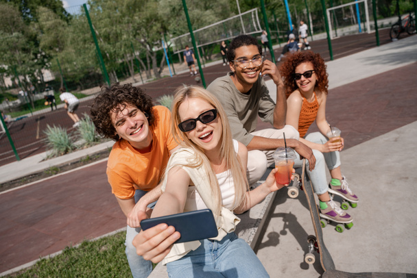 jovem feliz em óculos de sol tomando selfie com patinadores multiétnicos ao ar livre - Foto, Imagem