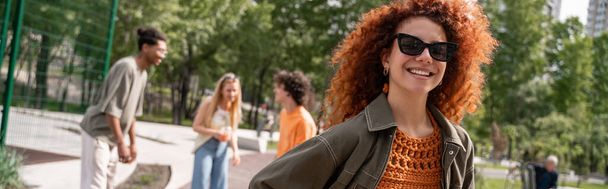 happy curly woman in sunglasses looking at camera near blurred interracial friends, banner - Foto, Imagen