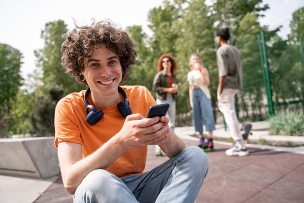 kudrnatý muž s mobilním telefonem s úsměvem na kameru v blízkosti rozmazané multietnické bruslaře - Fotografie, Obrázek