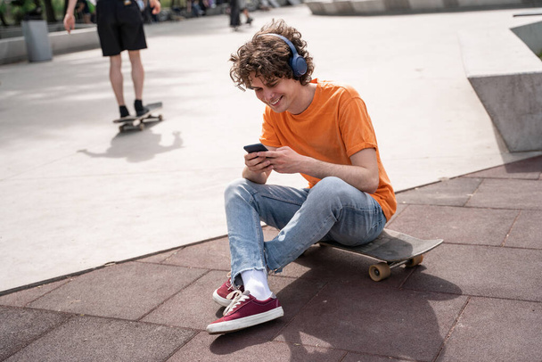 homme heureux dans les écouteurs assis sur skateboard et en utilisant smartphone dans le skate park - Photo, image