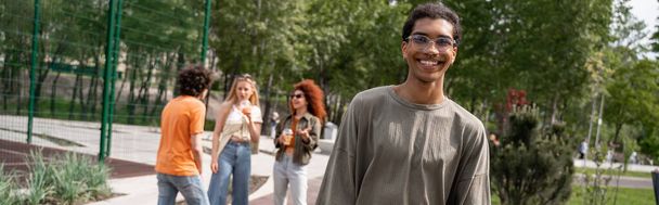 hombre afroamericano feliz mirando la cámara cerca de amigos en el parque, pancarta - Foto, Imagen