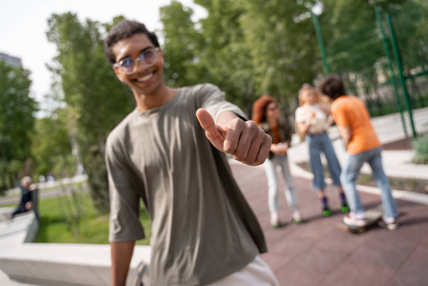 blurred african american man showing thumb up near blurred skaters in park - Foto, Imagen