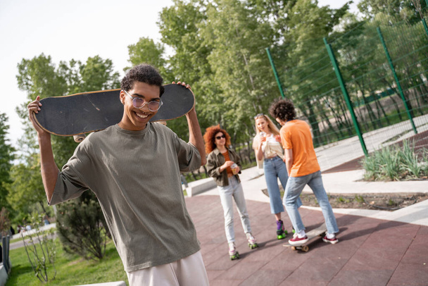 feliz homem americano africano com skate olhando para a câmera perto de amigos multiétnicos - Foto, Imagem