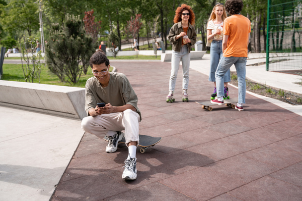 Χαρούμενος Αφροαμερικάνος που κουβεντιάζει στο κινητό του κοντά σε φίλους στο πάρκο για σκέιτ - Φωτογραφία, εικόνα