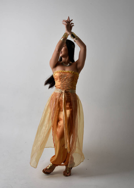 Ολο το μήκος πορτρέτο μιας όμορφης νεαρής Ασιάτισσας που φοράει χρυσές αραβικές ρόμπες σαν τζίνι, στέκεται με την πλάτη στην κάμερα, απομονωμένη στο φόντο του στούντιο. - Φωτογραφία, εικόνα