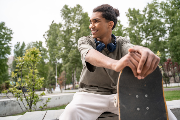 young and joyful african american skater looking away while sitting in skate park - Foto, Bild