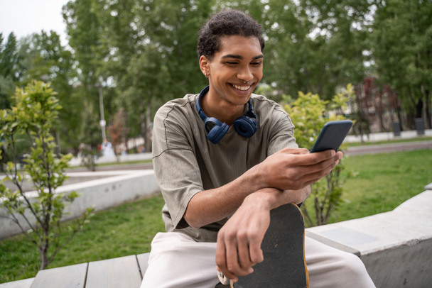 радісний афроамериканський фігурист сидить на прикордонній лавці і обмін повідомленнями на смартфоні
  - Фото, зображення