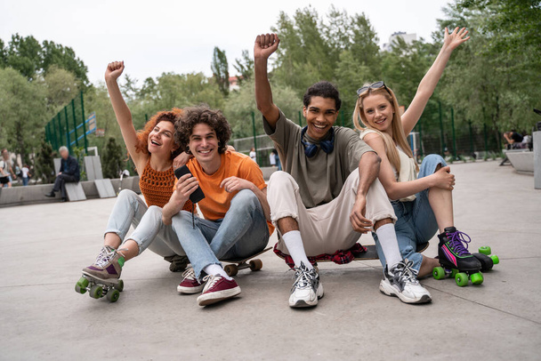 excited multicultural skaters showing win gesture while sitting on road in skate park - Photo, Image