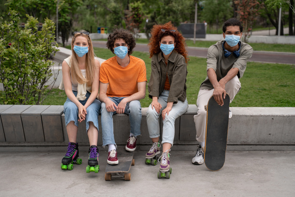 スケートパークの国境のベンチに座っている医療マスクの多民族の友人 - 写真・画像