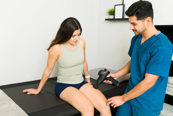 Relaxa. Fisioterapeuta profissional hispânico massageando a coxa de uma paciente caucasiana com uma arma de massagem  - Foto, Imagem
