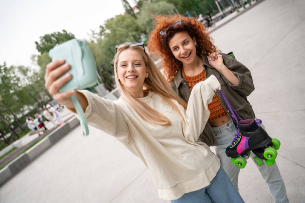 kihara nainen rullaluistimet hymyilee lähellä ystävä ottaa selfie digitaalikameralla - Valokuva, kuva