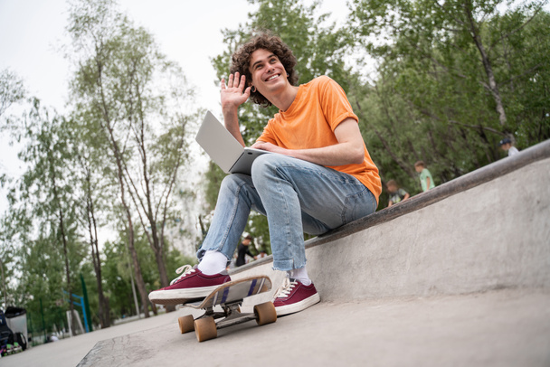 cheerful skateboarder with laptop waving hand while sitting outdoors - Fotoğraf, Görsel