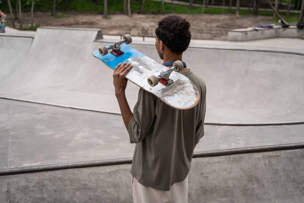 Африканский американец со скейтбордом на плече, смотрящий на скейт-рамп  - Фото, изображение