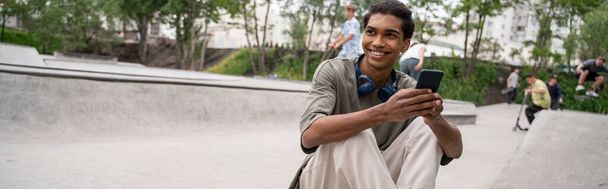スケートパークに座っているヘッドフォンや携帯電話を持つ幸せなアフリカ系アメリカ人男性 - 写真・画像