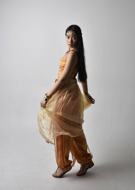 Retrato de larga duración de una hermosa joven asiática vestida con túnicas árabes doradas como un genio, posando de pie con la espalda a la cámara, aislada en el fondo del estudio. - Foto, imagen