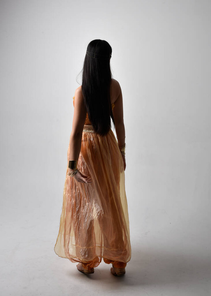 Retrato de larga duración de una hermosa joven asiática vestida con túnicas árabes doradas como un genio, posando de pie con la espalda a la cámara, aislada en el fondo del estudio. - Foto, imagen