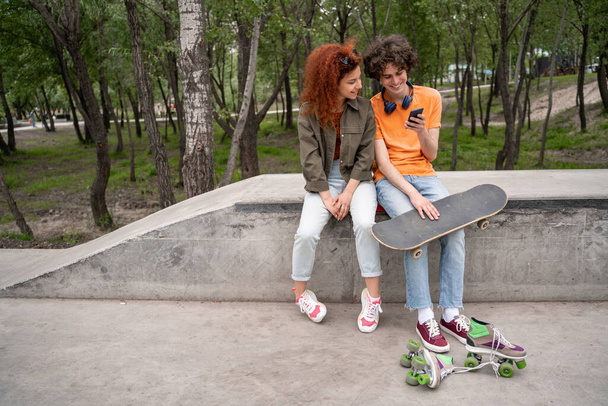 jong schaatser met behulp van smartphone terwijl zitten met een vriend op de grens in het park - Foto, afbeelding