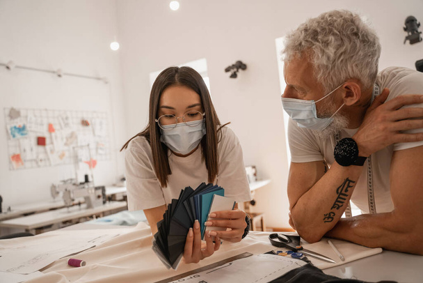 Asian designer in medical mask holding color swatches near colleague  - Photo, Image