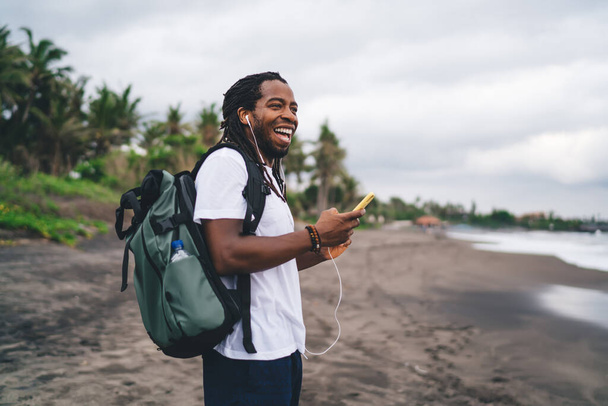 Side view joyful African American male in casual shirt using mobile phone and earphones while standing with backpack on exotic beach and looking away happily - Foto, afbeelding
