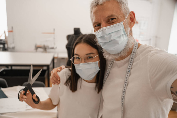 Diseñador en máscara médica mirando cámara cerca asiático colega con tijeras  - Foto, Imagen