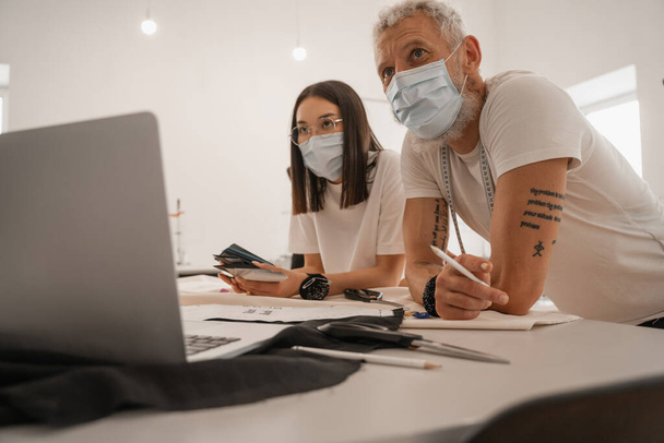 Fajok közötti tervezők orvosi maszkokban, homályos laptop közelében dolgoznak  - Fotó, kép
