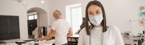Diseñador asiático en máscara médica mirando a la cámara mientras colega borrosa trabajando en el estudio, pancarta  - Foto, Imagen