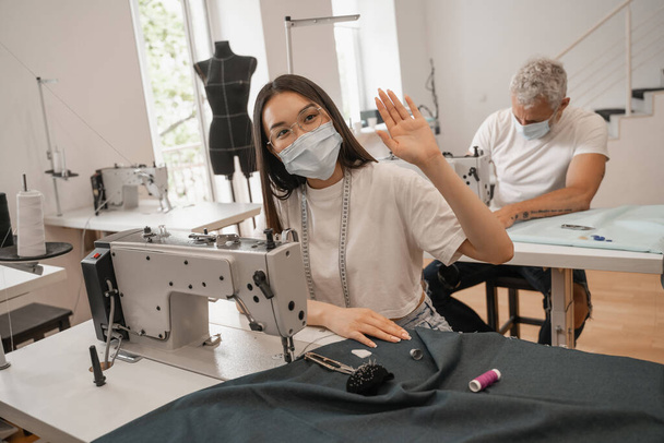 Costurera asiática en máscara médica agitando la mano cerca de la máquina de coser y colega  - Foto, Imagen
