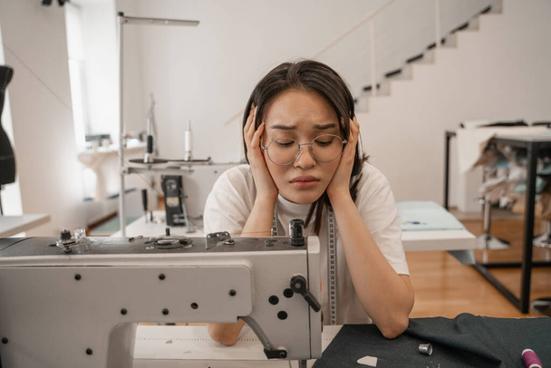 Tired asian seamstress looking at sewing machine and cloth  - Zdjęcie, obraz