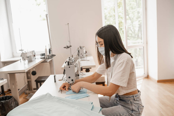 Asian designer in medical mask sewing in atelier  - 写真・画像