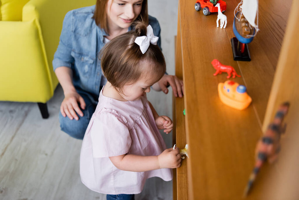 Down sendromlu küçük bir kız, gülümseyen anaokulu öğretmeninin yanındaki tahta rafta oyuncaklara bakıyor. - Fotoğraf, Görsel