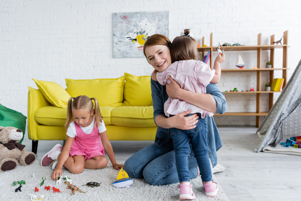 счастливая воспитательница детского сада обнимает малышку с синдромом Дауна рядом с дошкольником, играющим с игрушками в игровой комнате  - Фото, изображение