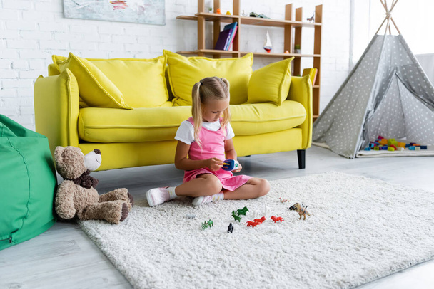Vorschulmädchen sitzt auf Teppich und spielt mit Spielzeug im modernen Spielzimmer  - Foto, Bild
