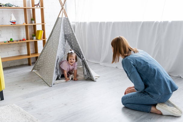 kindergarten teacher looking at happy kid with down syndrome sitting in tipi - Fotó, kép