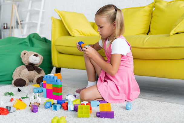 blonde girl in pink dress playing building blocks on carpet in kindergarten playroom  - 写真・画像