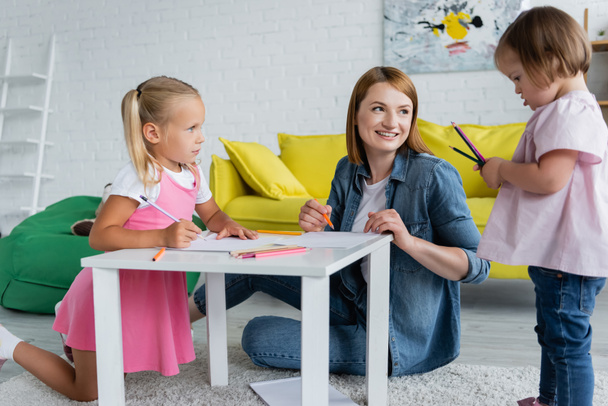 улыбающаяся воспитательница детского сада и дошкольница смотрит на ребенка с синдромом Дауна, держащего цветные карандаши  - Фото, изображение