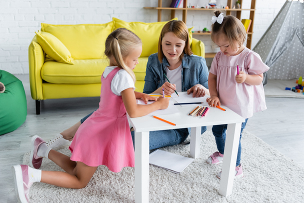 улыбающийся воспитатель детского сада разговаривает с дошкольником и ребенком с синдромом Дауна рядом с бумагами и цветными карандашами  - Фото, изображение