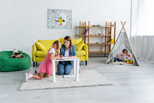 kindergarten teacher looking at girl drawing while toddler kid with down syndrome sitting in teepee  - Foto, Bild