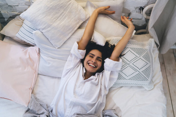 Joyful female in sleepwear stretching while awaking during positive sunday morning, happy Caucasian woman in glad mood awaking during lazy day on weekend relaxing in comfortable home bedroom - Valokuva, kuva