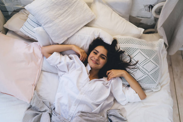 Top portrait view of carefree female feeling peaceful pleasure during sunday morning and long lying in bed, happy Caucasian woman relaxing during lazy weekend day looking at camera after wake up - Zdjęcie, obraz
