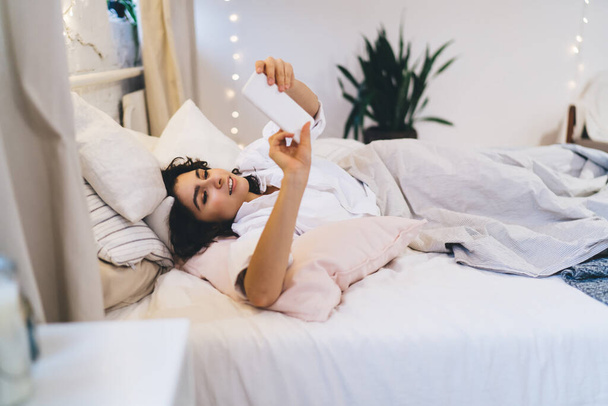 Beautiful woman checking network news after awaking using smartphone gadget and home internet connecting while lying in comfortable bed, Caucasian female blogger sending message via mobile app - Zdjęcie, obraz