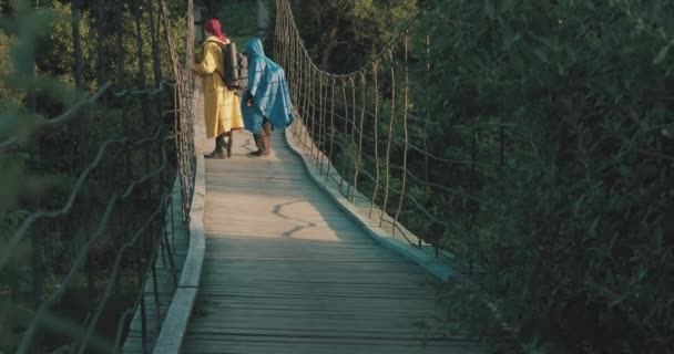 Caminantes hombres y mujeres en impermeables caminan en puente colgante. Puesta de sol - Imágenes, Vídeo