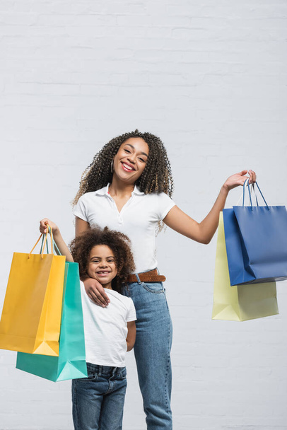 alegre mujer afroamericana con niño sosteniendo coloridas bolsas de compras en gris - Foto, imagen