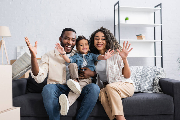 joyful african american family waving hands on couch in new apartment - Photo, Image