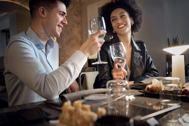 Счастливая межрасовая пара веселится, разговаривая, обедая и выпивая в ресторане Fusion Food. Молодые люди встречаются с звенящими бокалами. - Фото, изображение