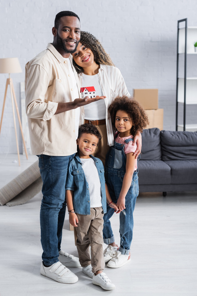 happy african american man holding house model near wife and children in new apartment - Fotó, kép