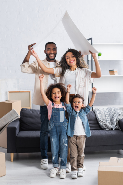 enfants afro-américains excités montrant geste de succès près des parents avec toit en carton dans un nouvel appartement - Photo, image