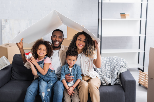 fröhliches afrikanisch-amerikanisches Paar mit Kindern auf Sofa unter Papierdach - Foto, Bild