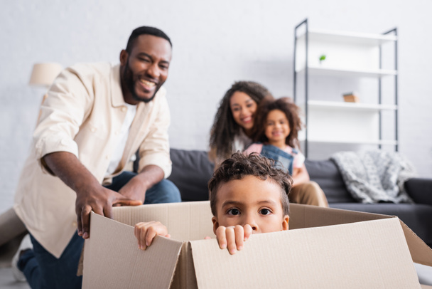 niño afroamericano sentado en caja de cartón cerca de la familia alegre sobre fondo borroso - Foto, imagen