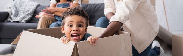 nadšený africký americký chlapec sedí v kartonové krabici poblíž rozmazané rodiny, prapor - Fotografie, Obrázek